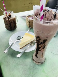 On a white plate, there is a slice of cheese pastry accompanied by spoons, and on the table, there are two chilled chocolate beverages.