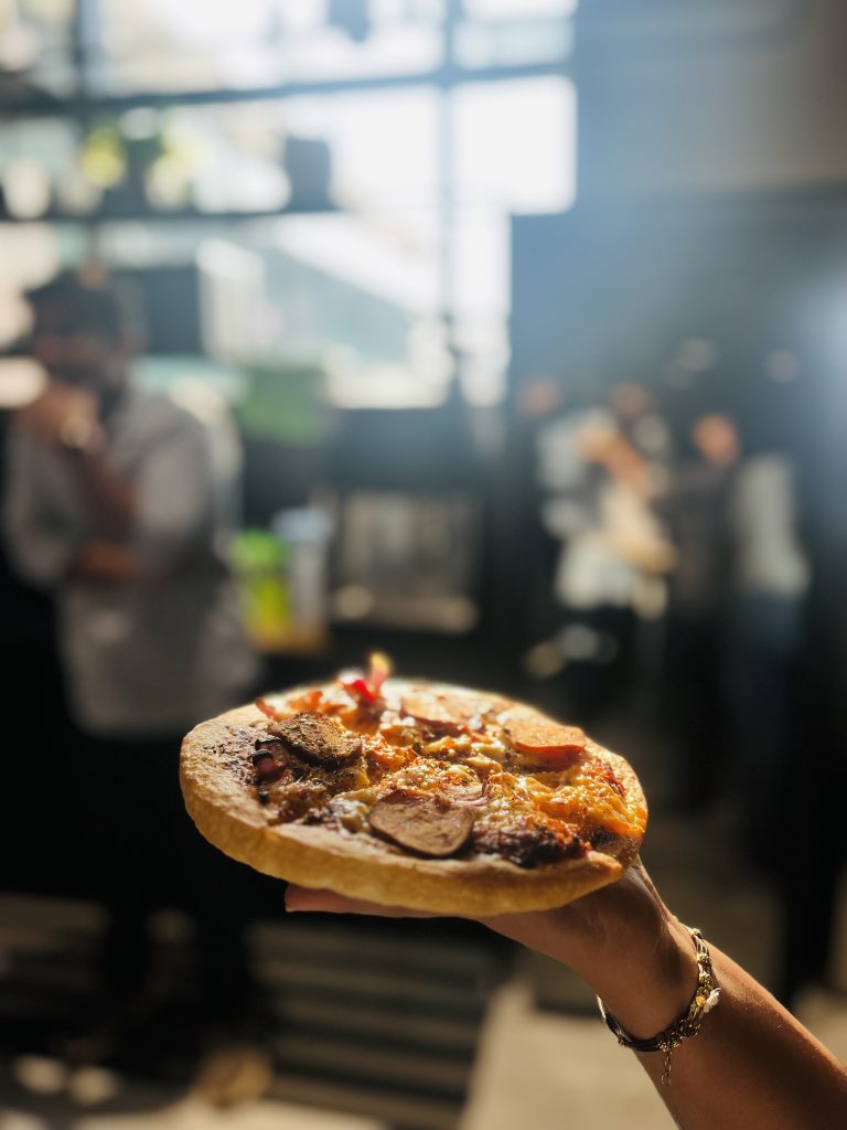 A person’s hand holding up a freshly made pizza with sausage and various toppings, in a blurry indoor setting with other people in the background.