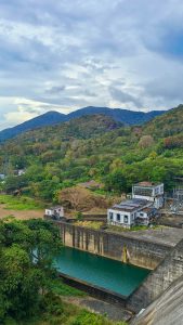 A picturesque scene unfolds at Thenmala Dam, where lush greenery, majestic mountains, and the gentle flow of water from the dam create a captivating view. 