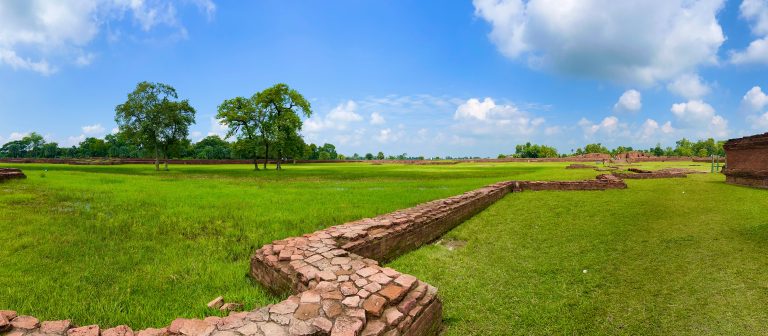 A panoramic view of a lush green field with ancient brick ruins in the foreground, scattered trees throughout, under a blue sky with scattered clouds.