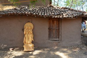 Golden statue of Hindu Lord Radhe Krishna, stands on dusty, dry soil, leaning against a wooden door house in a Rajasthan village, it's roof heavy with tiles secured in part by string. To the right a man comes into view.