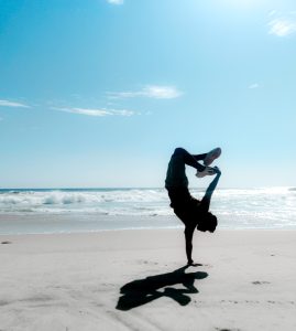 A sillhouette of a person doing a one-hand handstand on the beach. Waves approach from behind.