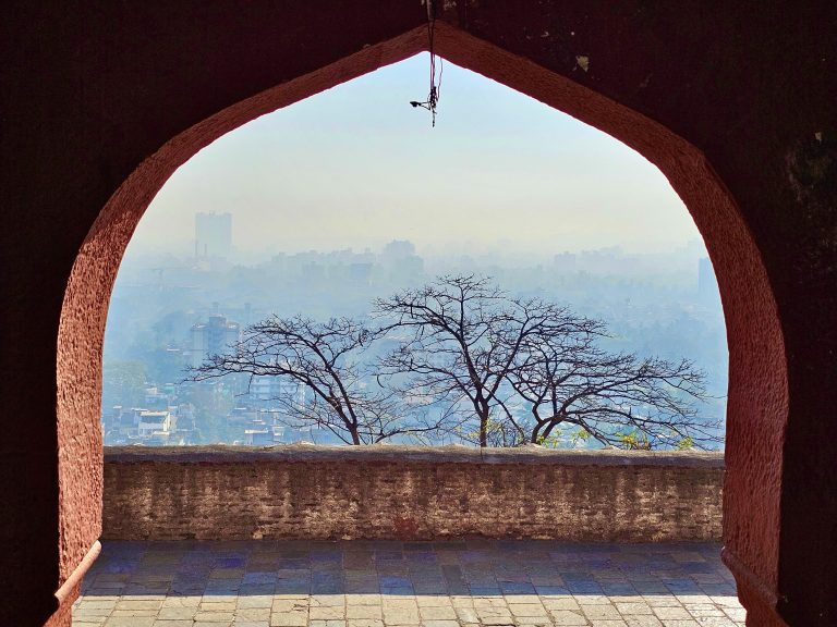 An early morning view from an arched doorway, with leafless trees silhouetted against the city and a hazy sky in the background. Parvati hills, Pune, Maharashtra.