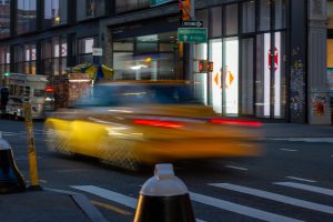 一辆黄色出租车在纽约市的一条街道上快速行驶，背景是食品车和一家银行。
