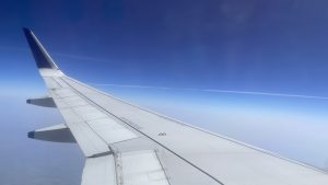 A white and grey airplane wing on the left side of the plane against a blue sky with contrail.
