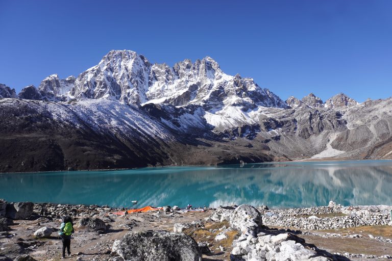 People are standing on the bank of the lake with snow covered mountain in the background on a clear sunny day. Gokyo Lake