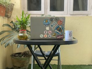 Laptop adorned with vibrant stickers illustrating a WordPress enthusiast’s journey, on a table with a lucky bamboo plant and a mug on each side. Each sticker tells a story of achievements and community love.
