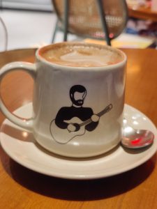 A cappuccino is a coffee concoction that consists of espresso, served in a white ceramic cup and saucer, adorned with an illustration of a man playing a guitar.