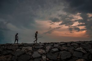  Silhouettes of two people walking on rocky terrain against a dramatic sky at sunset.