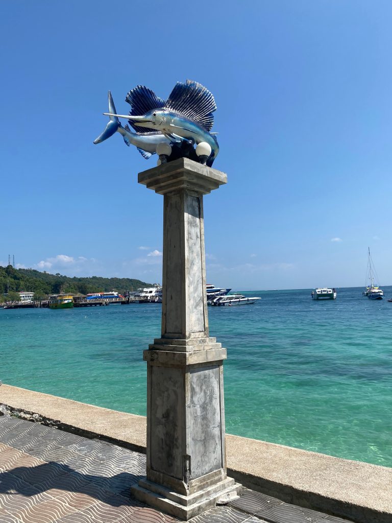 A street light towers over whimsical fish sculptures, framed by the azure sea and bustling cruise boats, a charming blend of art and coastal life.