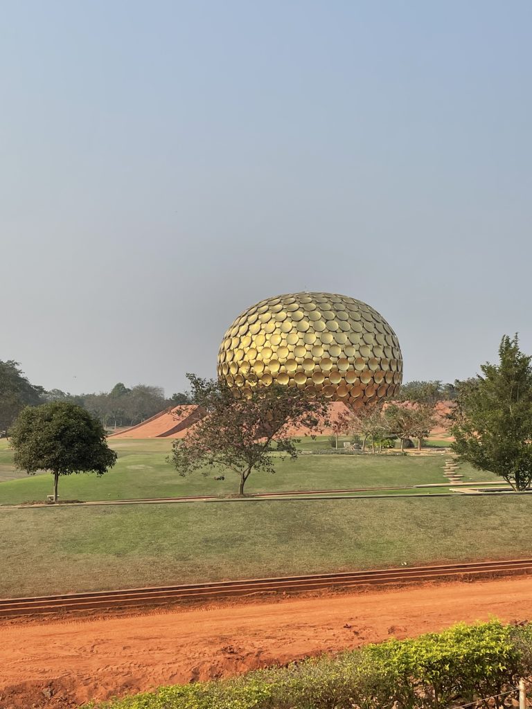 Matrimandir a golden sphere structure constructed from circle and triangle shapes, edged with brick slopes in Auroville, Pondicherry stands in a grass park dotted with trees.