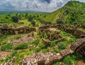 An aerial view of Bhangarh Fort, Rajasthan amidst green vegetation, with a mountainous backdrop and a cloudy sky.