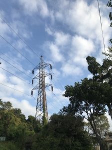 wires passing along the transmission tower, with trees in the foreground, under a cloudy, blue sky. 