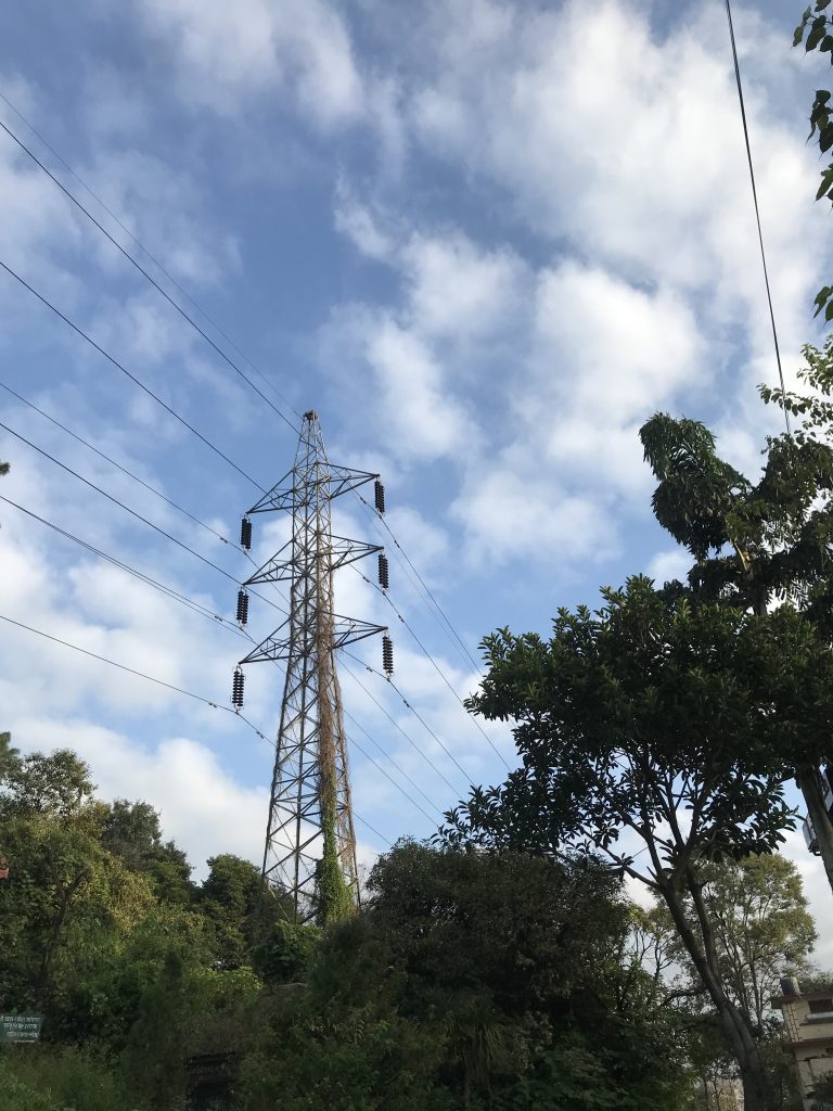 wires passing along the transmission tower, with trees in the foreground, under a cloudy, blue sky.