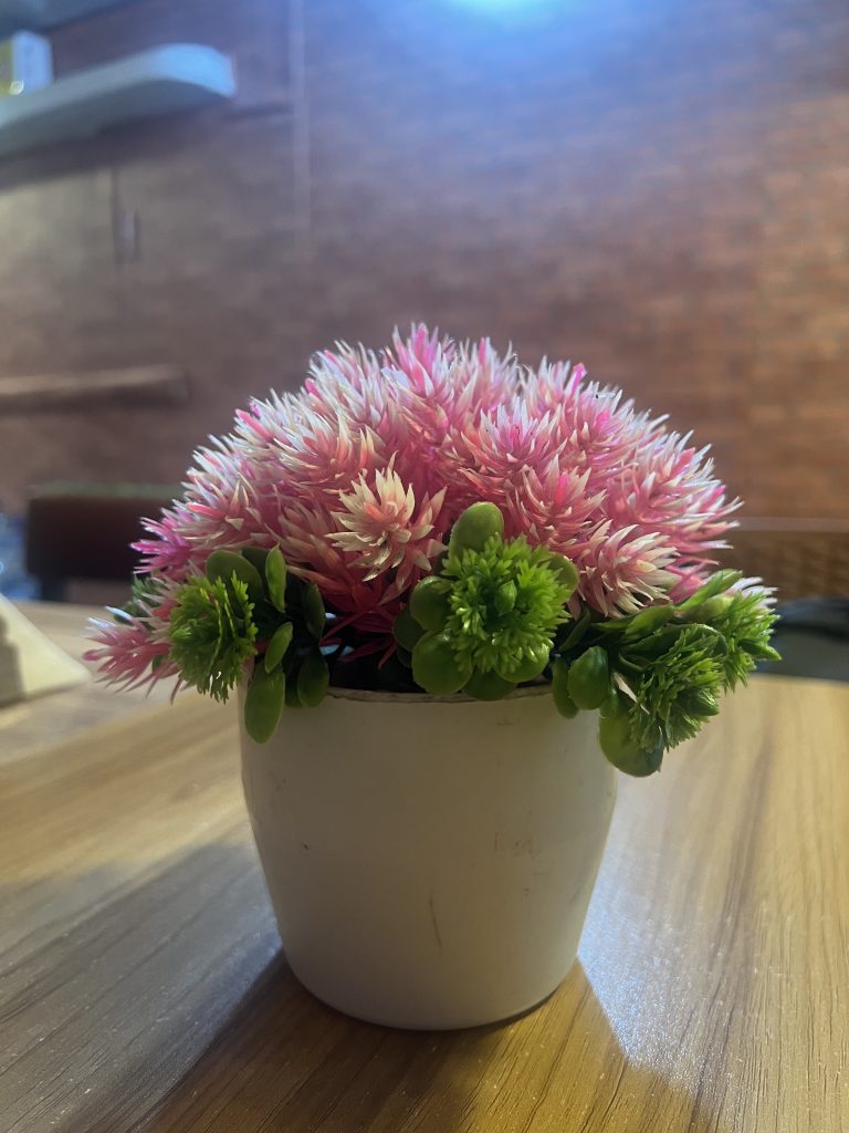 A pink plastic flower in a white flowerpot sitting on a wooden table.