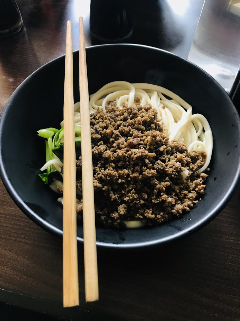 Noodles with minced meat in a dark bowl alongside chopsticks.