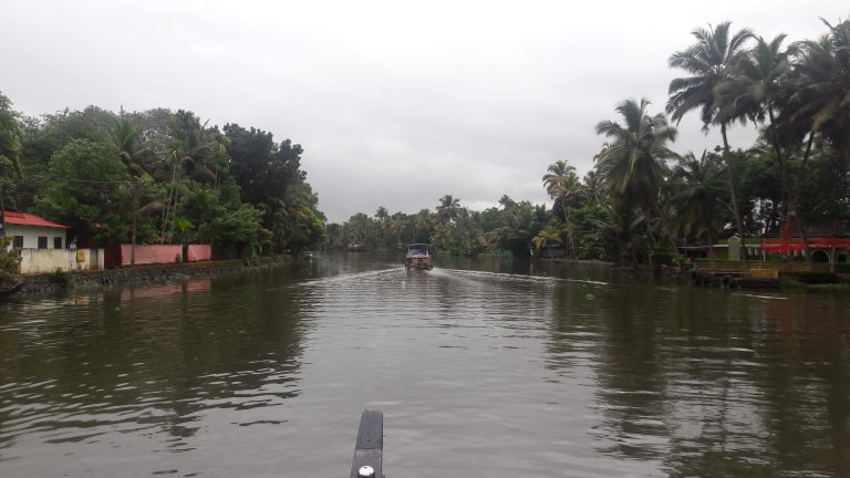 A view from a river looking at a boat ahead, and buildings and trees on the banks