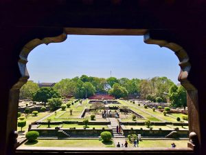 A view of a landscaped garden with symmetrical pathways, hedges, and trees, framed through an arch, with people walking and a clear blue sky above. Shaniwarwada, Located in Pune, Maharashtra. 