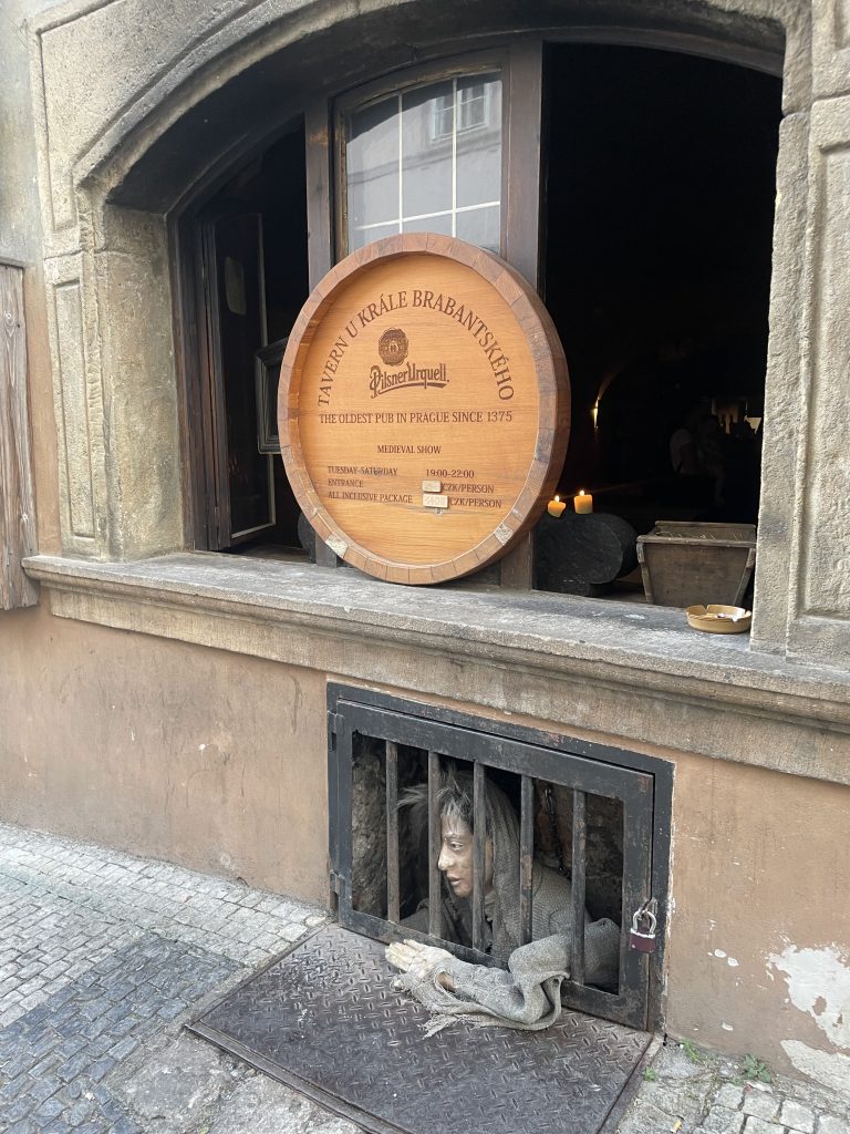 Exterior of the oldest pub in Prague, looking into the window. A mannequin reaches out at street level between bars in a window.