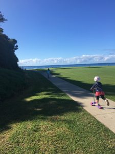 一个戴着头盔和安全装备的孩子在阳光明媚的公园里的水泥路上骑着踏板车，背景是草地，可以看到大海和天空。
