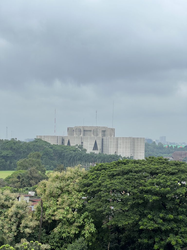 A grey geometric building rises above trees in front of a city