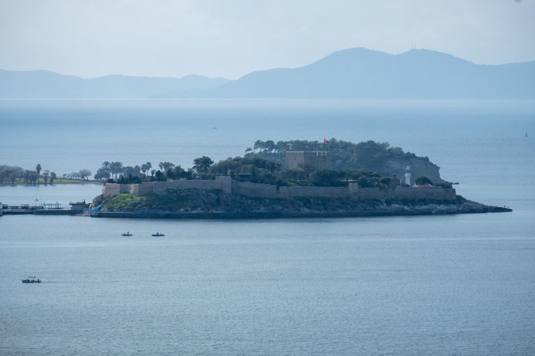 A photo of the Güvercinada shot from a distance. On the foreground, there are three small boats. Silhouette of Samos is seen in the background.