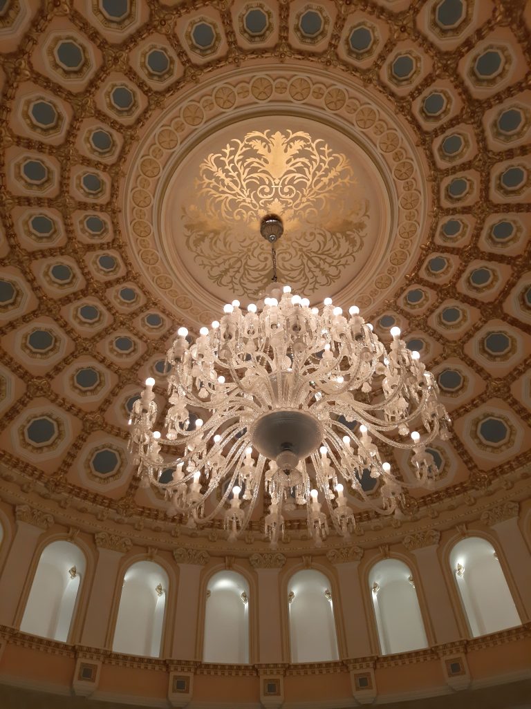 Grand chandelier with numerous lights hanging from an ornate ceiling with decorative molding and a circular pattern design.