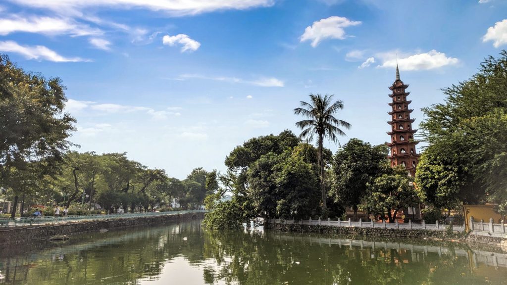 Tran Quoc Pagoda in Hanoi, Vietnam. The pagoda is situated in the trees beside a river with blue skies and clouds overhead.