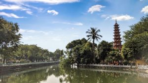 Tran Quoc Pagoda in Hanoi, Vietnam. The pagoda is situated in the trees beside a river with blue skies and clouds overhead.
