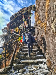 A person climbing a narrow staircase beside prayer flags while hiking, carrying a bag, with a cloudy sky in the background. 