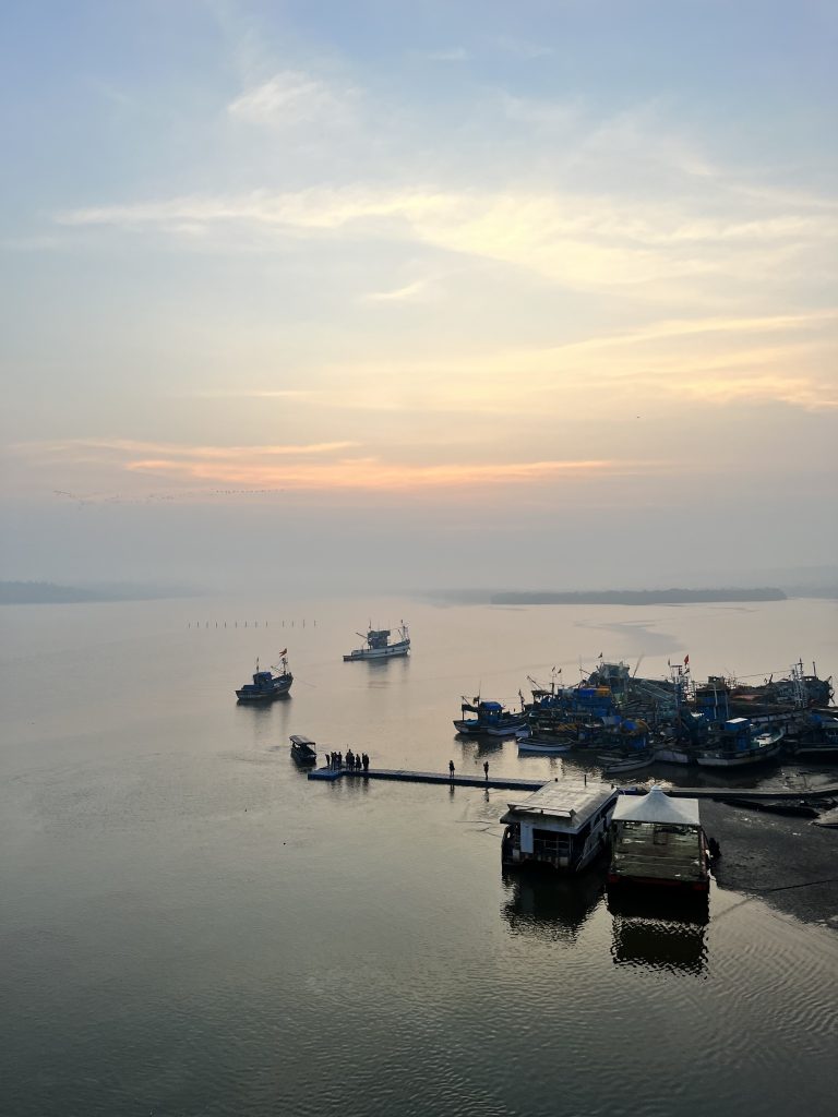 Sunrise over a large expanse of water in Goa. Small boats moored together at the shore, larger boats moored in the water. People on the pier looking out at the view.