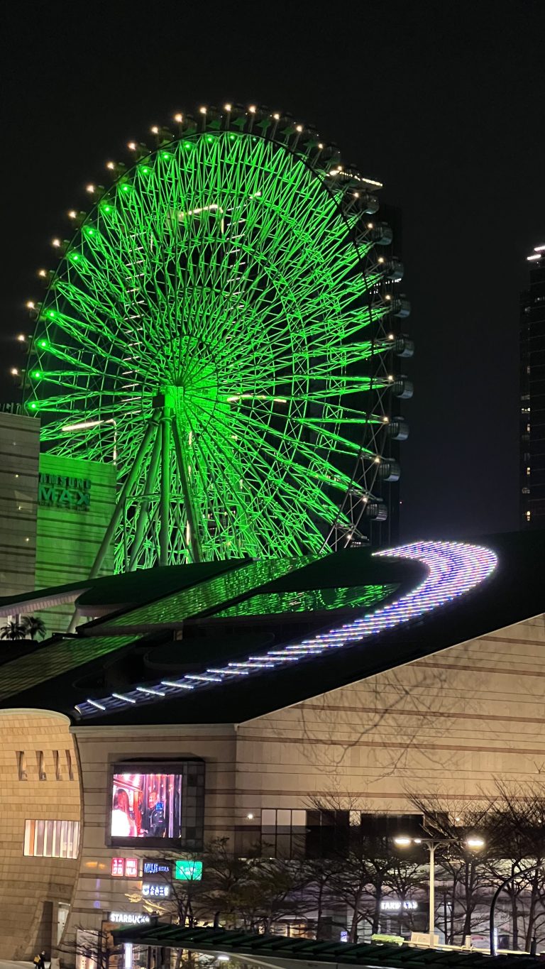 A brightly lit Ferris wheel with green illumination at night, surrounded by urban buildings and a walkway featuring blue light accents.