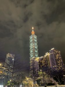A night view of the brightly lit Taipei 101 towering over surrounding buildings against a cloudy night sky.