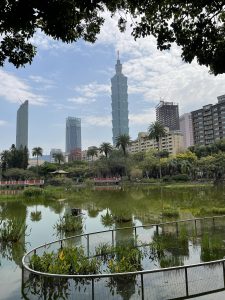 A cityscape featuring a prominent, tiered skyscraper (Taipei 101) and surrounding buildings as seen from Zhongshan Park in Taipei, Taiwan with lush greenery and a pond in the foreground. Leaves from overhanging trees frame the top of the image.