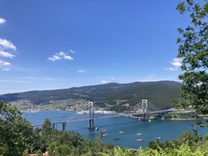 A view of a picturesque Puente de Rande en la Ria de Vigo featuring a large cable-stayed bridge spanning a blue body of water with floating platforms, surrounded by lush green hills and a clear sky with a few clouds.