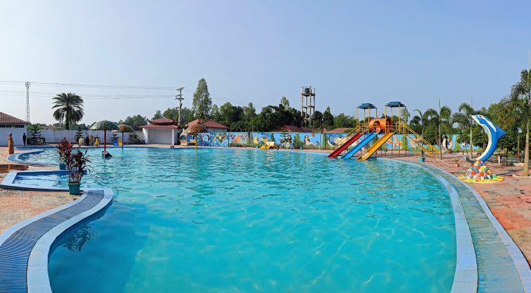 A panoramic view of a large outdoor swimming pool with colorful water slides and artistic murals on the surrounding walls under a clear blue sky.