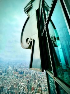 View of Taipei from the window of the observation floor of the Taipei 101 building, with its iconic embellishment in the foreground and large reflective glass panels.
