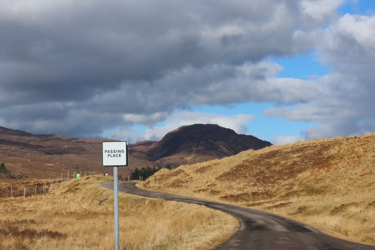 Scottish Highland winding single track road with passing place sign. Mountain lies ahead, dry dead grasses cover the land each side of the road.