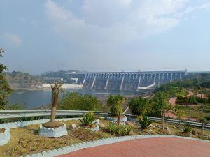 A panoramic view of Sardar Sarovar Dam with spillways, a reservoir, surrounding hills, and a landscaped park area with pathways and tropical plants in the foreground.
