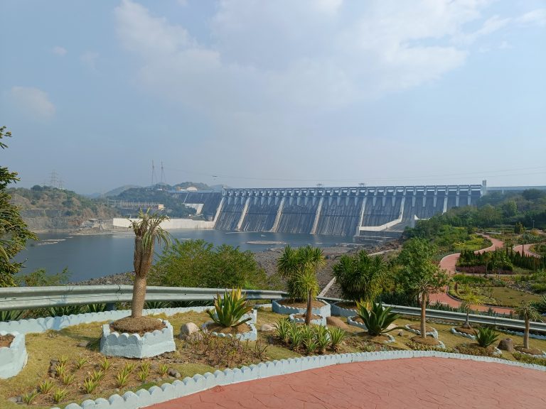 A panoramic view of Sardar Sarovar Dam with spillways, a reservoir, surrounding hills, and a landscaped park area with pathways and tropical plants in the foreground.