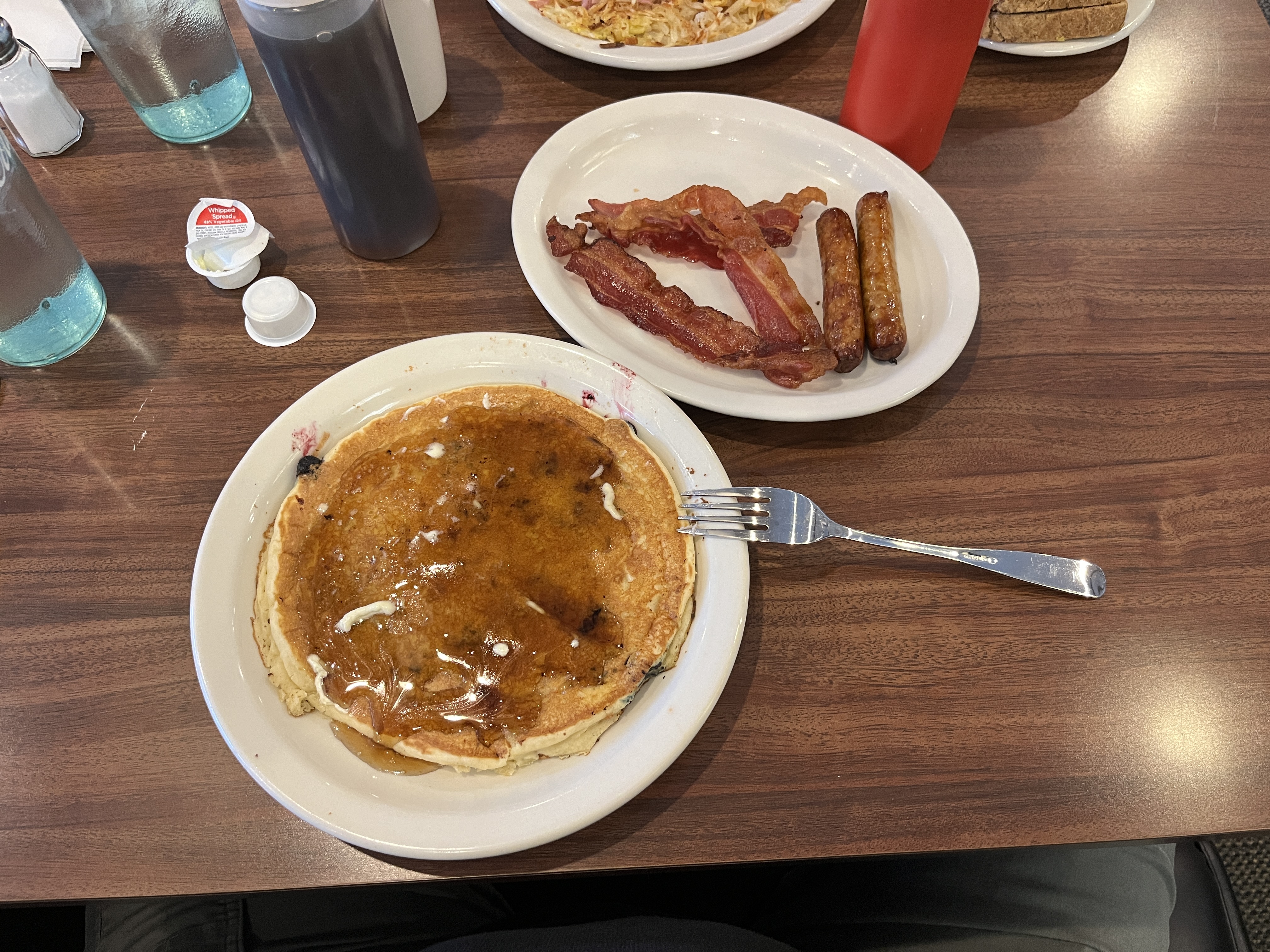 Fork rests on a plate of pancakes covered in syrup and another plate of bacon rashers and sausages sit on a formica table with surrounded by condiments, water, napkins and a used spread mini pots. Barely in shot two more plates of food, looks like a sandwich and an omelette,