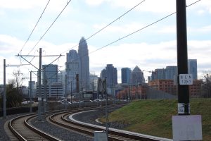 The skyline of the city of Charlotte, North Carolina seen through the rail lines of public transportation. There are buildings, cars, a railroad track and a mound with grass.

