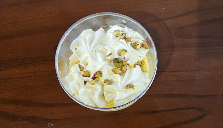 A dessert in a plastic bowl topped with whipped cream and pistachios, placed on a wooden table.