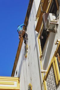 A Painter in a blue shirt and protective gear is suspended by ropes while repairing or painting the exterior of a building.

