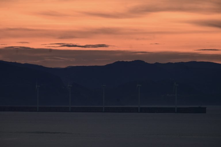 A serene sunset at the dock, where the sky is painted in vivid hues of orange, pink, and purple as the sun dips below the horizon, wind turbines stand tall, their blades turning slowly in the gentle breeze, generating clean electricity.