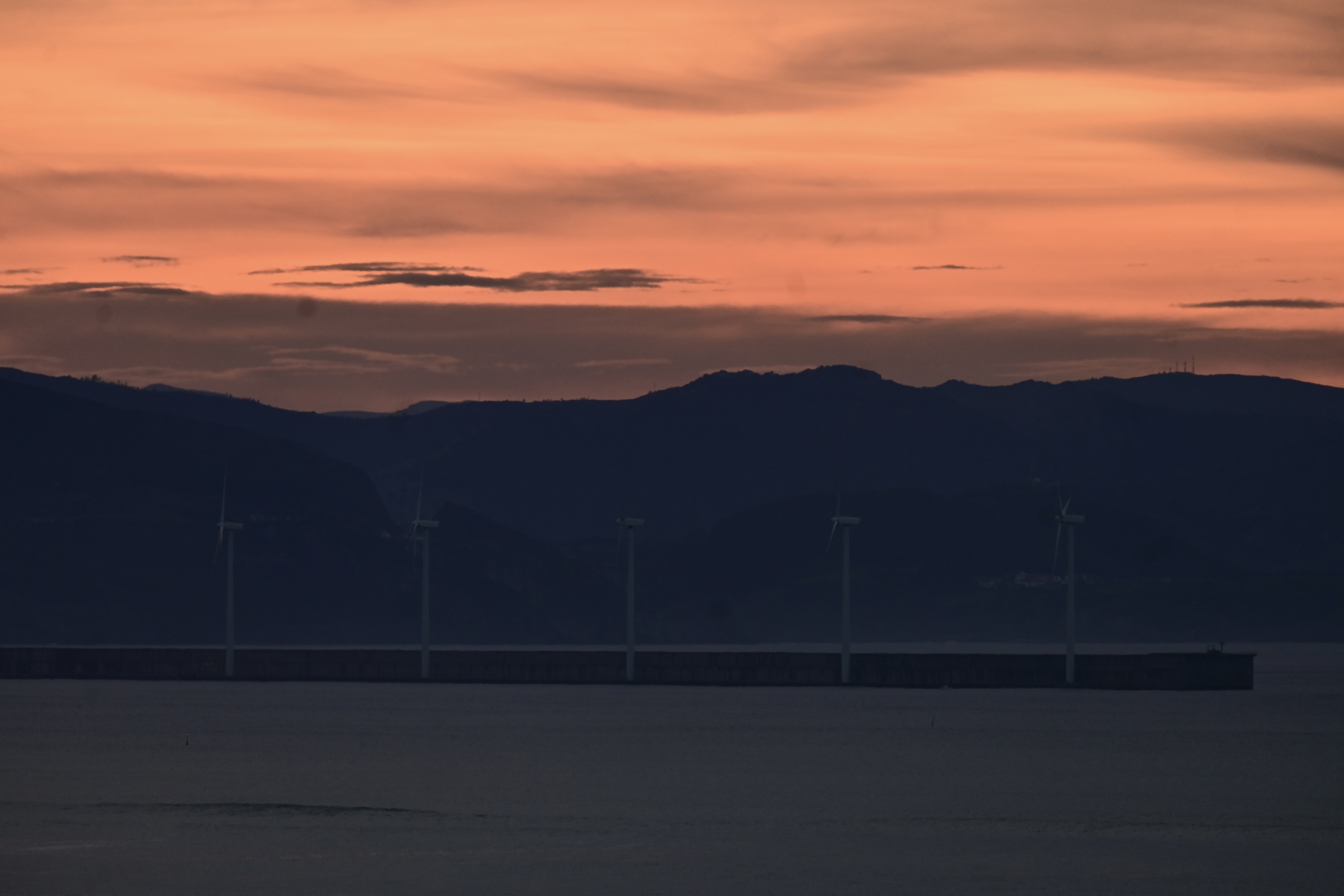 A serene sunset at the dock, where the sky is painted in vivid hues of orange, pink, and purple as the sun dips below the horizon, wind turbines stand tall, their blades turning slowly in the gentle breeze, generating clean electricity.