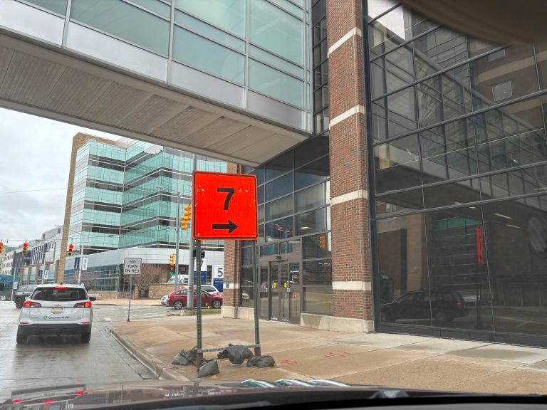 Orange construction sign with a 7 on it under a walking bridge by a building and a road.