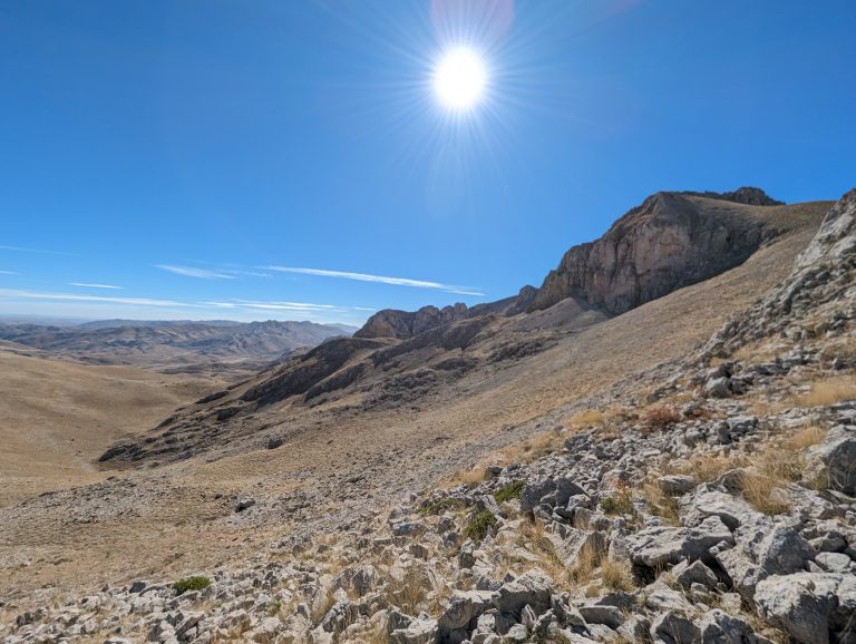 A sunny landscape featuring dry, rocky terrain with large mountains in the background under a bright blue sky with a prominent sun. Geyik mountains, Turkey.