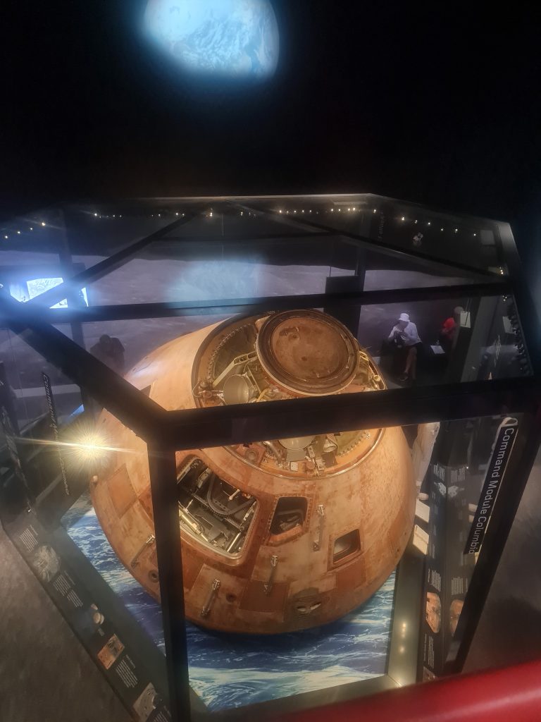 The photo shows an overhead view of a historic space capsule with its hatch open, displayed in a museum setting, with a backdrop featuring a large image of Earth as seen from space