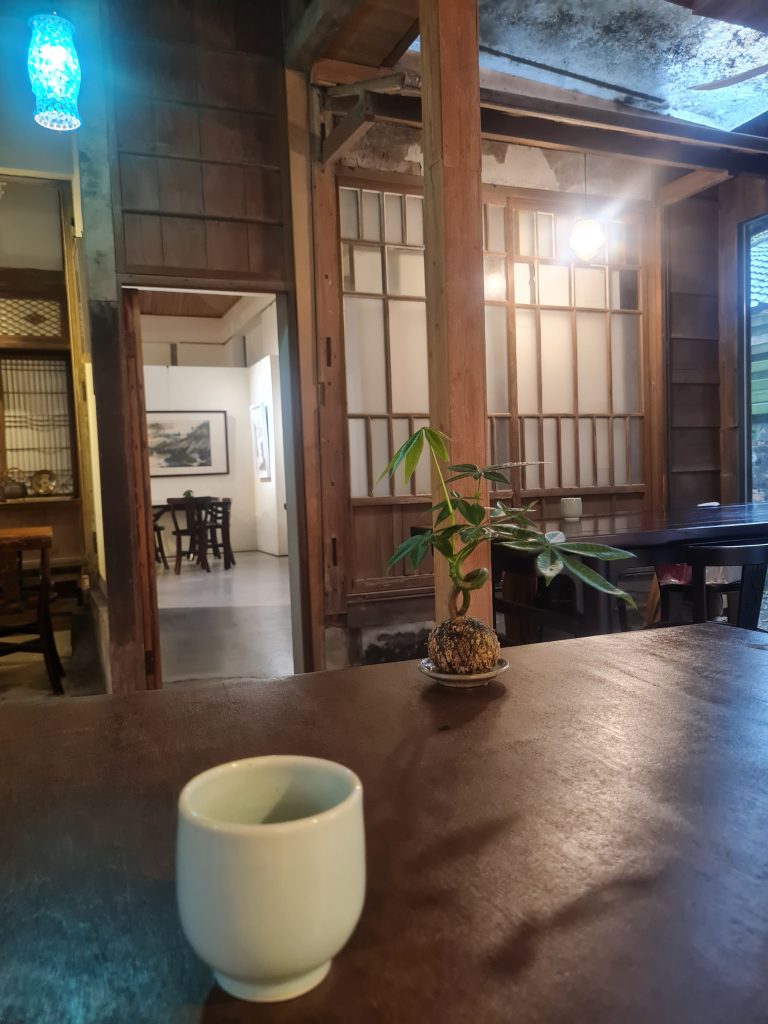 This image shows a serene interior, likely of a traditional Japanese house, with a focus on a small cup and a potted plant on a table, and featuring sliding doors and hanging lanterns in the background.